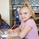 a child sitting at a desk and holding a pencil smiles at the camera